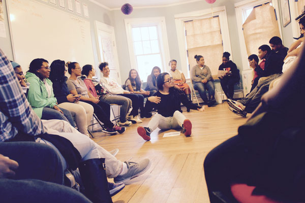 A large group of youth sitting in a circle with one person demonstrating on the floor in the middle.