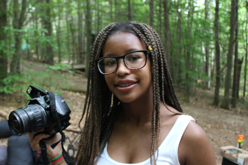 A young person with long braids standing amongst green trees holding a professional digital camera