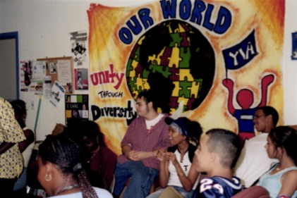 A small group of teenagers laughs while looking at something off-camera. A banner hangs on the wall behind them with an image of the earth made of puzzle pieces, reading "OUR WORLD", "YIA", and "UNITY THROUGH DIVERSITY".