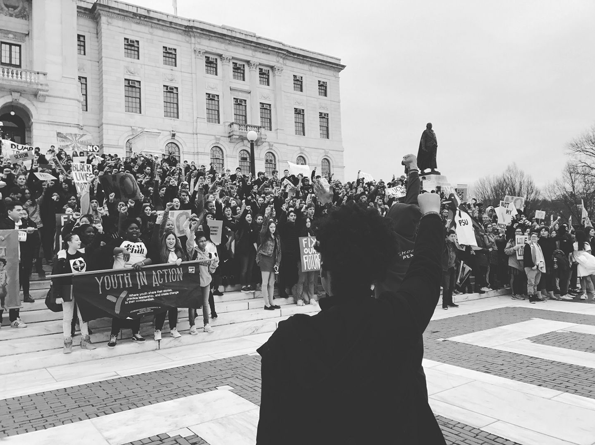 A large crowd of youth stand in front of the Rhode Island state house with fists raised in the air, some holding a banner or protest signs.