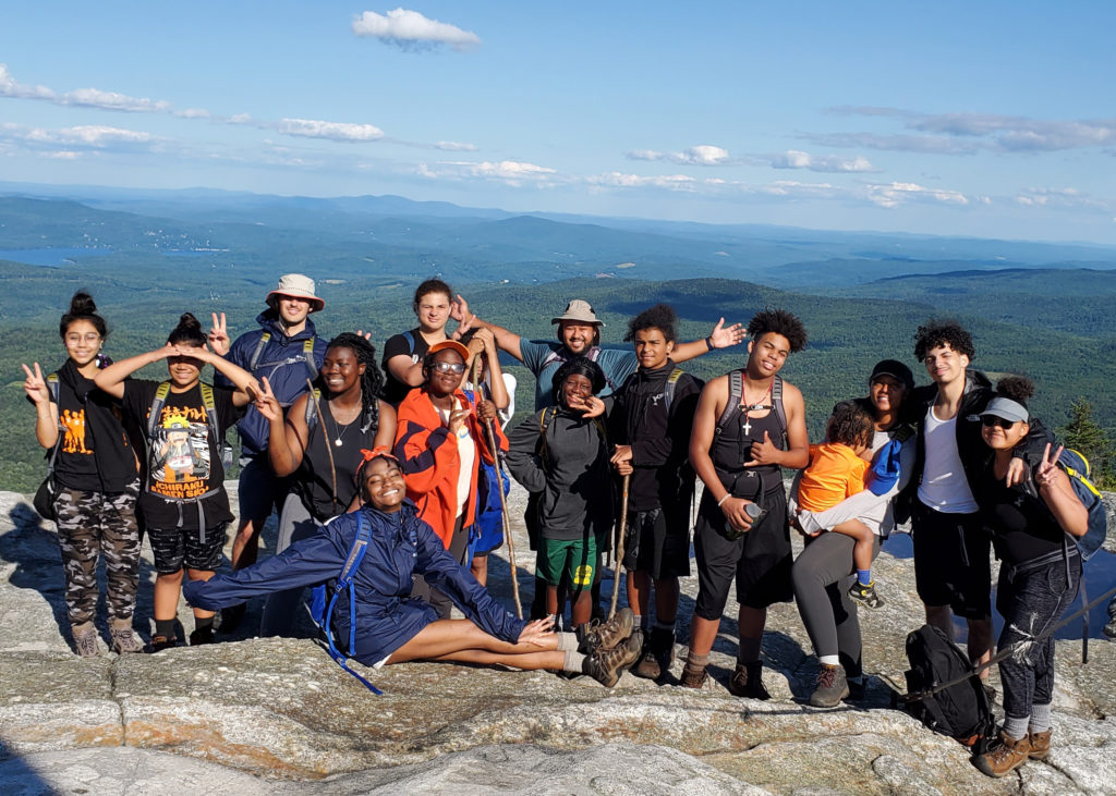 A smiling group of youth, a couple adults, and a child holding each other and giving peace signs atop a rocky summit overlooking a blue sky and rolling hills.