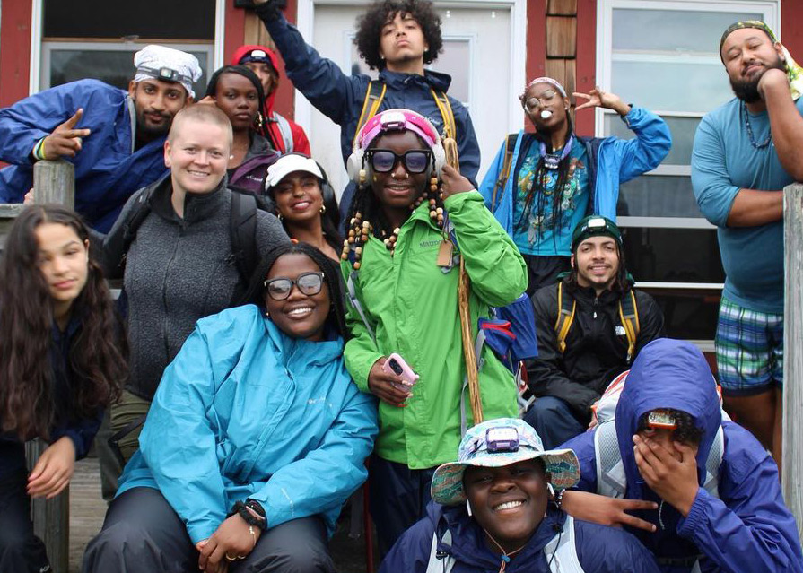 About a dozen youth and a couple adults smiling and wearing rain gear pose in front of a red cabin.