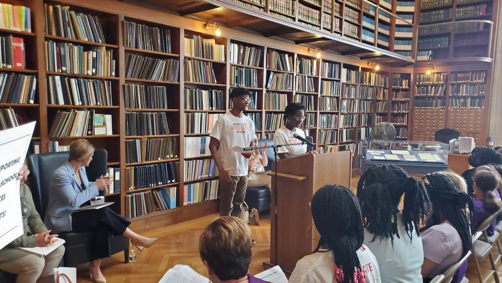 Two youth stand at a podium in front of an audience, with two stories of bookshelves behind them.