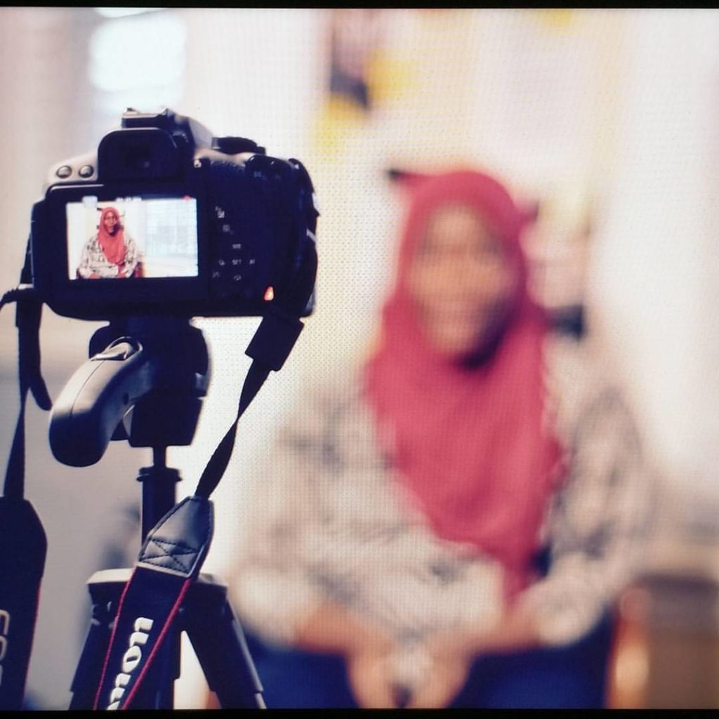 A camera on a tripod records a young person in hijab sitting.