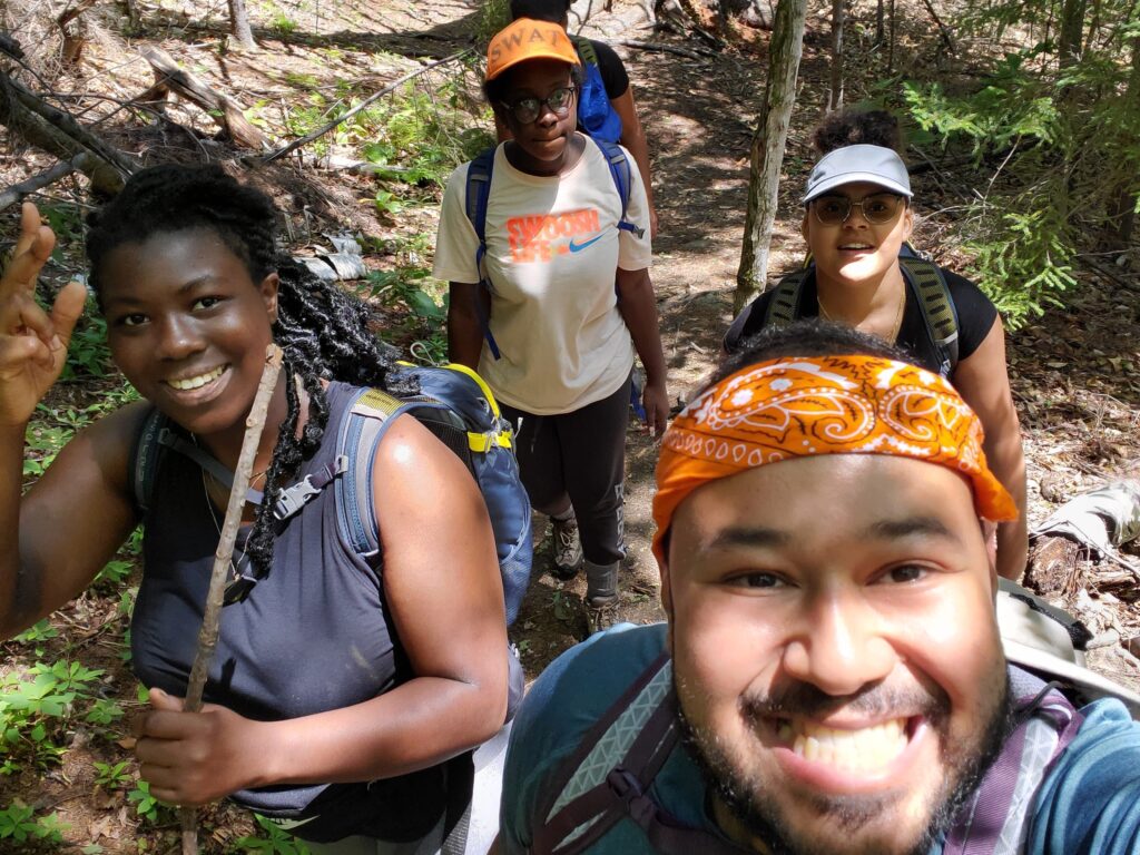 Four people in the woods smile at the camera, wearing camping gear and backpacks.