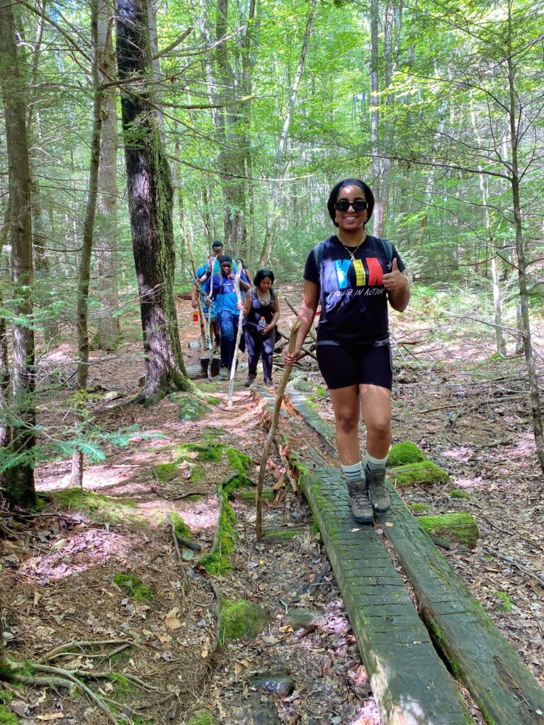 Person gives a thumbs up walking along a forest path, a few others follow behind with walking sticks.